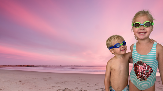 Boy wearing blue Frogglez swimming goggles looks up at his older sister who is earing green swim goggles as they stand on a beach with a pink sunset