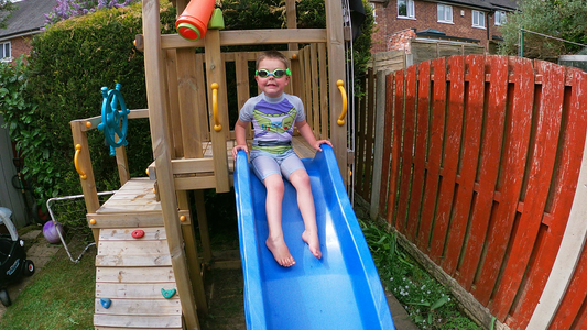 Cute boy wearing Frogglez on backyard slide into pool. Get Frogglez in the UK at Swim Buddies. Frogglez available on Amazon. Boys swim goggles. 