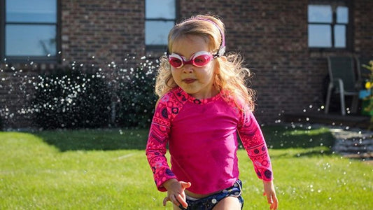 Little girl wearing pink mermaid Frogglez swim goggles with blue swim diaper and pink rash guard. Best goggles for girls. Running through sprinklers.