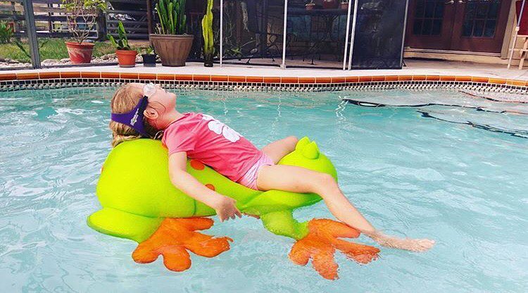 Girl swimmer in backyard swimming pool floating on a frog in a pink rashguard wearing Frogglez swim goggles. Frogglez goggles have a sturdy headband strap that is easy to use. Adjust easily with velcro.