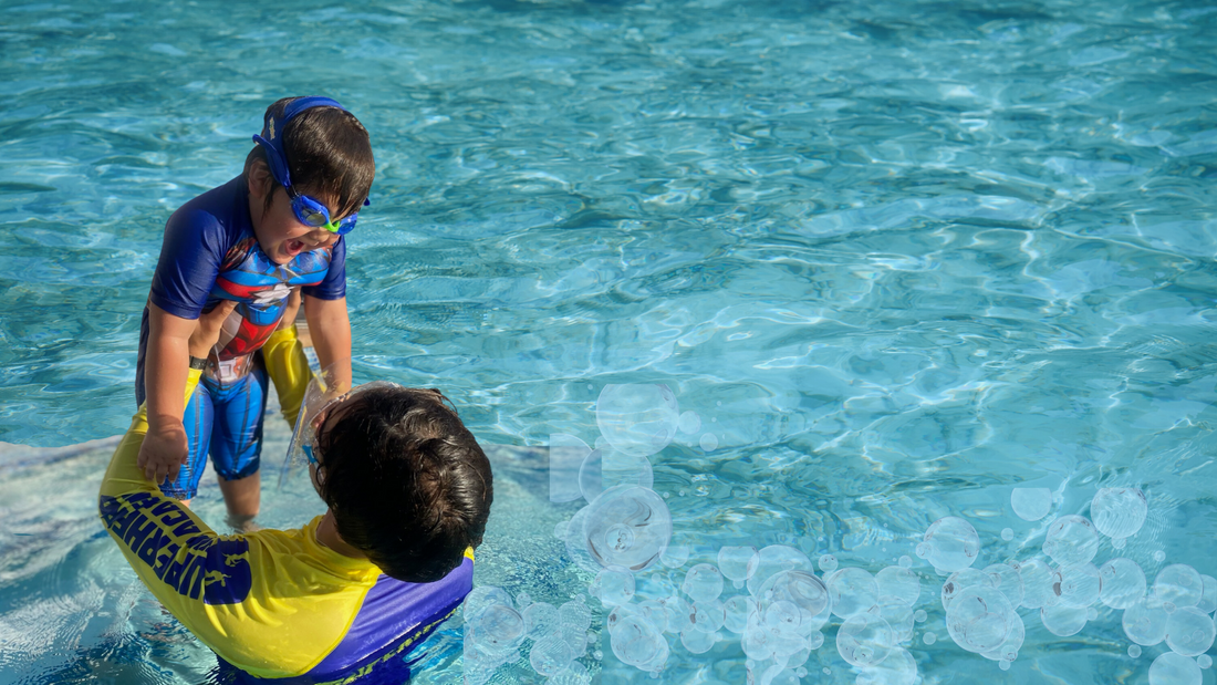 Swim instructor holds a laughing child just above the water in a swimming pool during swim lessons. Boy is wearing rashguard and blue Frogglez Goggles 