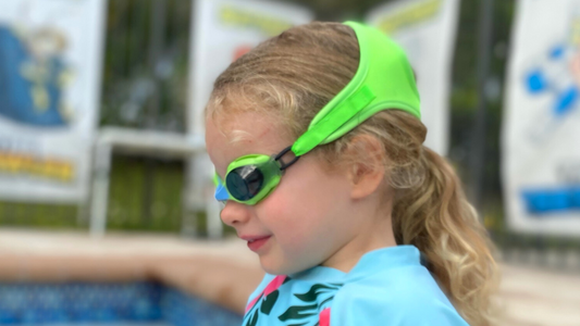 Girl sitting on swimming pool steps wearing green Frogglez swim goggles. Frogglez comfortable neoprene strap stretches and won't pull hair. Best swim goggles for boys. Best swim goggles for girls.