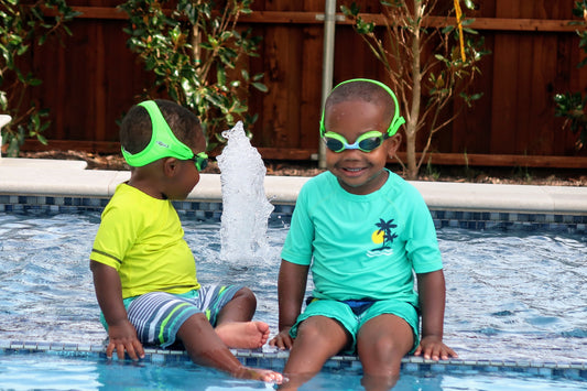two young boys in swim suits and swim shirts sit in the pool while wearing green Frogglez Kids Swim Goggles.