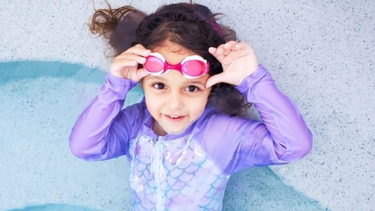 girl with pink frogglez goggles in a purple swimsuit lays down on swimming pool steps. froggles are the best kids swimming goggles. Kids should wear swim goggles during swim lessons to protect their eyes. 