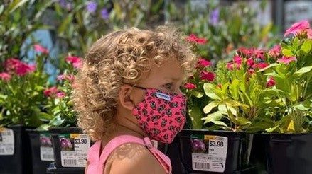 cute girl with curly hair wearing Frogglez pink cloth face mask with frog print. In front of flowers outside in the sunshine. Comfortable contoured face mask made just for kids.