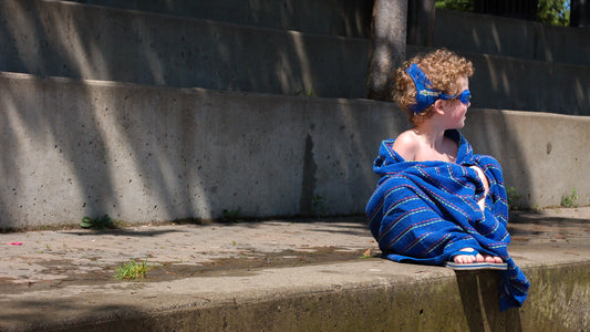 Boy with curly hair wearing Frogglez swim goggles sitting on the side of a swimming pool wrapped in a blue towel